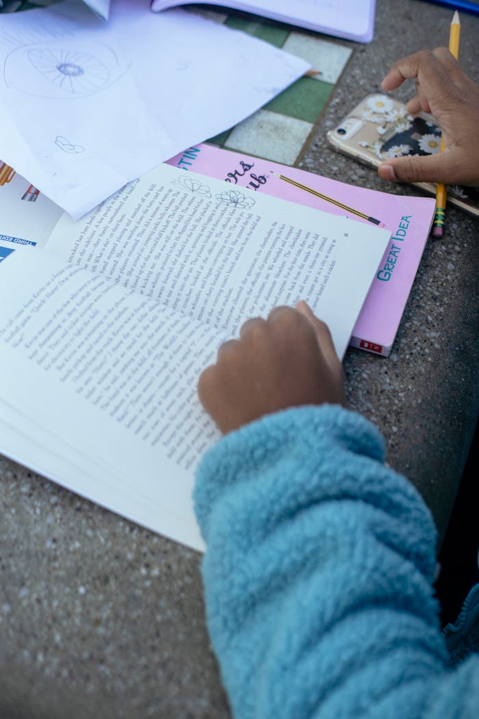 High angle of crop ethnic kid sitting at table with textbook and mobile phone while doing homework